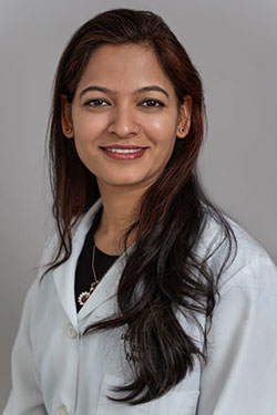 The image shows a woman with long hair, wearing a white lab coat, smiling at the camera. She has a professional appearance and is likely in a medical or healthcare setting.