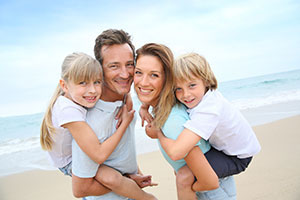 A family of four posing on a beach with the father holding the children and the mother in the background.