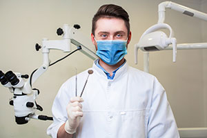 A man in a lab coat and face mask, holding a microscope slide, stands behind a dental chair with a microscope on a stand.