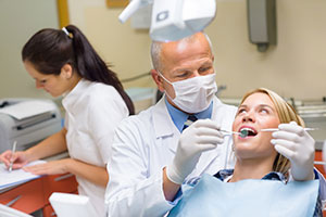 The image shows a dental office setting, with a dentist in white lab coat and mask examining a patient s mouth. There are other dental professionals present, including one woman wearing a blue surgical gown and another person in the background.