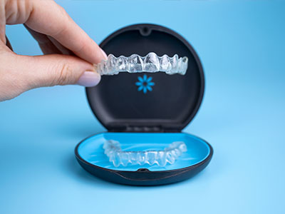 A human hand holding a clear plastic container displaying a set of transparent dental implants, with the brand name  Invisalign  visible on the top left corner.