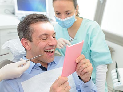 A man in a dental chair is holding up a card with his mouth, while a dentist and hygienist look on.