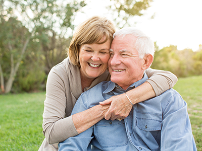 The image is a color photograph of an older man and woman embracing each other outdoors, with the woman wearing a light-colored top and the man in a dark shirt. They appear to be enjoying a moment together.