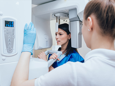 The image shows a woman in blue medical scrubs standing next to a large, modern 3D scanner with a digital display, while another person is seated and appears to be observing or interacting with the equipment.