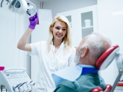 A dental hygienist is holding a mirror over an elderly man s face, both in medical gowns with masks and gloves.