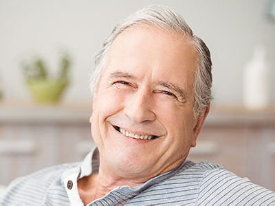 An elderly man with a smile, wearing glasses and sitting in an indoor setting.